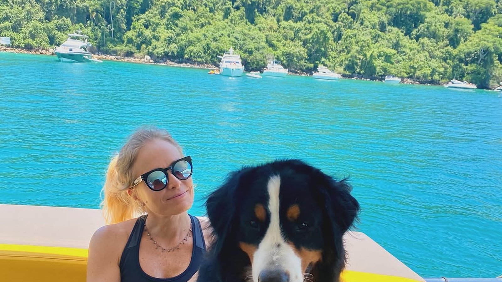 Angelica de oculos escuros sentada em um barco ao lado do cao Obama, com o mar azul ao fundo