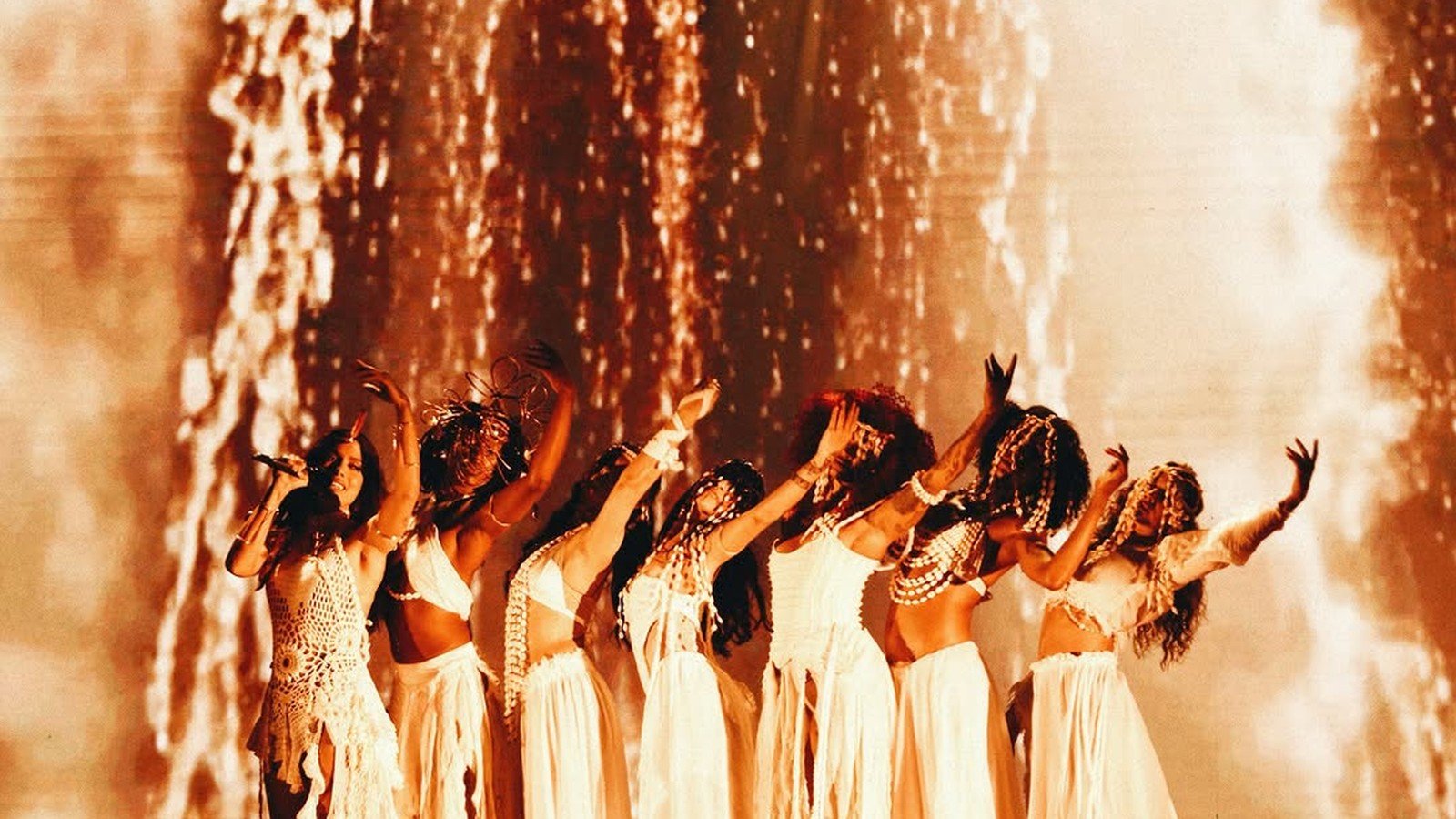 Bailarinas de branco enfileiradas no palco da turne da Anitta sob luz dourada