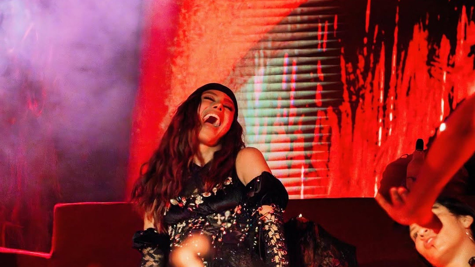 Anitta cantando de vestido preto brilhante sob luz vermelha no palco