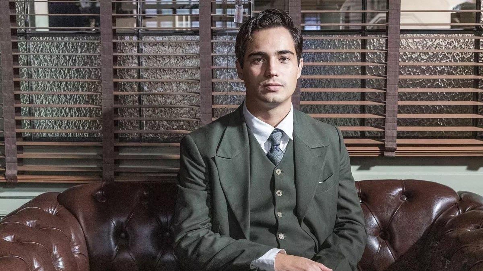 Danilo Mesquita sentado num sofá de couro, de terno verde com colete e gravata