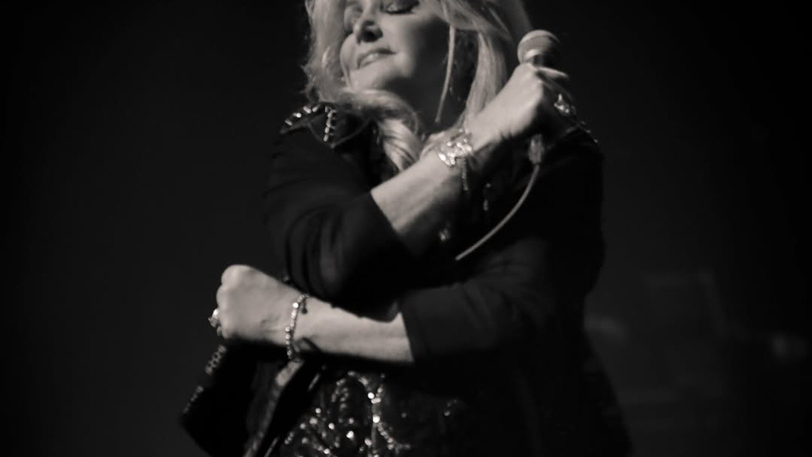 Bonnie Tyler cantando no palco de casaco preto, com os bracos cruzados sobre o microfone