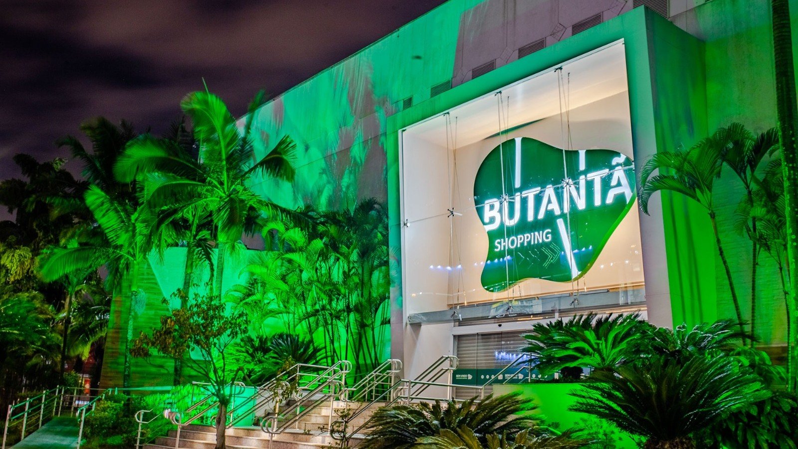 Fachada iluminada de verde do Butanta Shopping a noite, com palmeiras em primeiro plano