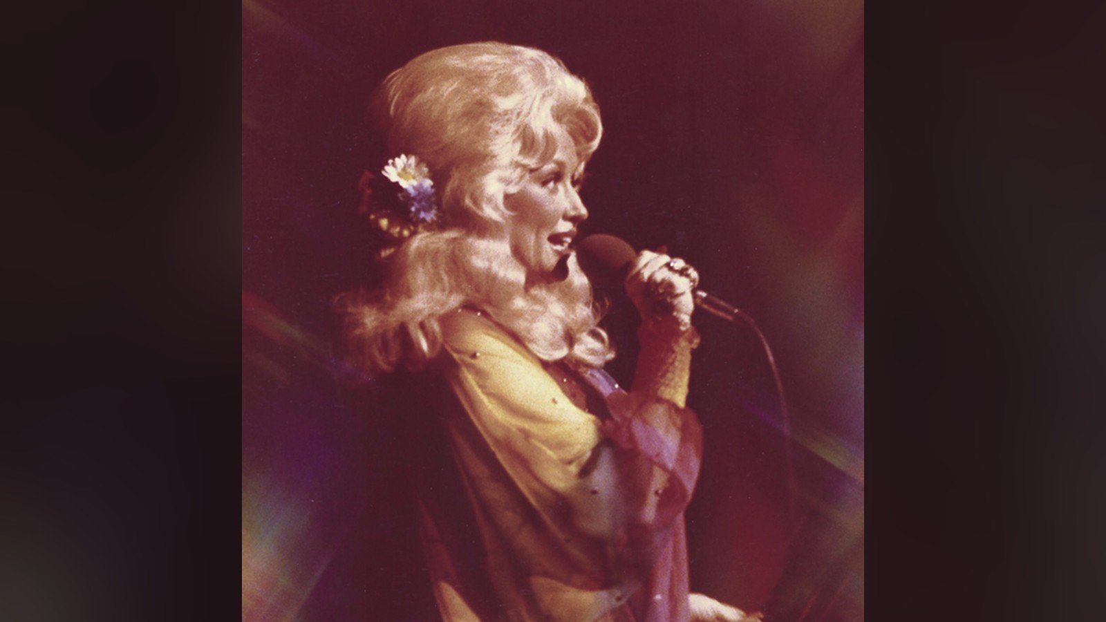 Dolly Parton cantando ao microfone, cabelo loiro volumoso, em show