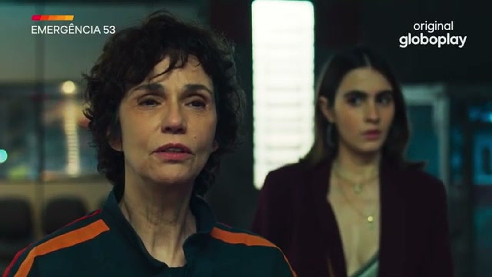 Yara de Novaes de uniforme do SMU ao lado de uma personagem de casaco vinho, na base da ambulancia