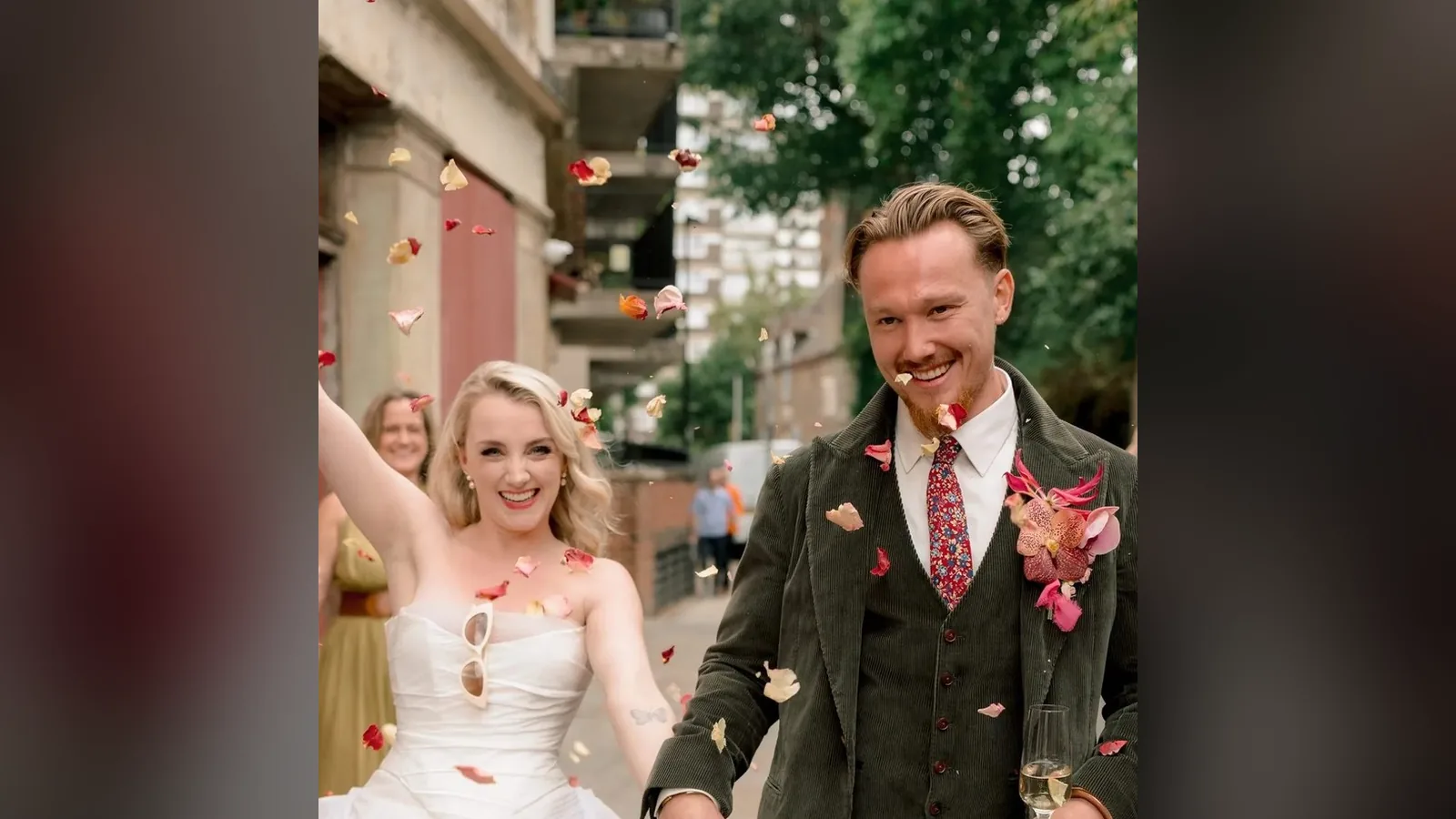 Evanna Lynch e Denis Skinner saem do casamento sob uma chuva de pétalas, ela de vestido branco e ele de terno verde