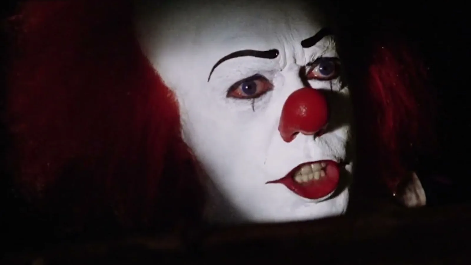Tim Curry como o palhaço Pennywise, de rosto branco, nariz vermelho e cabelo ruivo, dentro de um bueiro