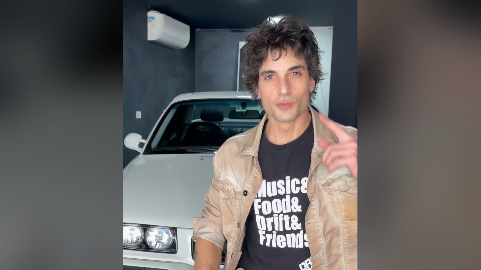 Fiuk sentado num banco, de jaqueta clara e camiseta preta, diante de um carro branco