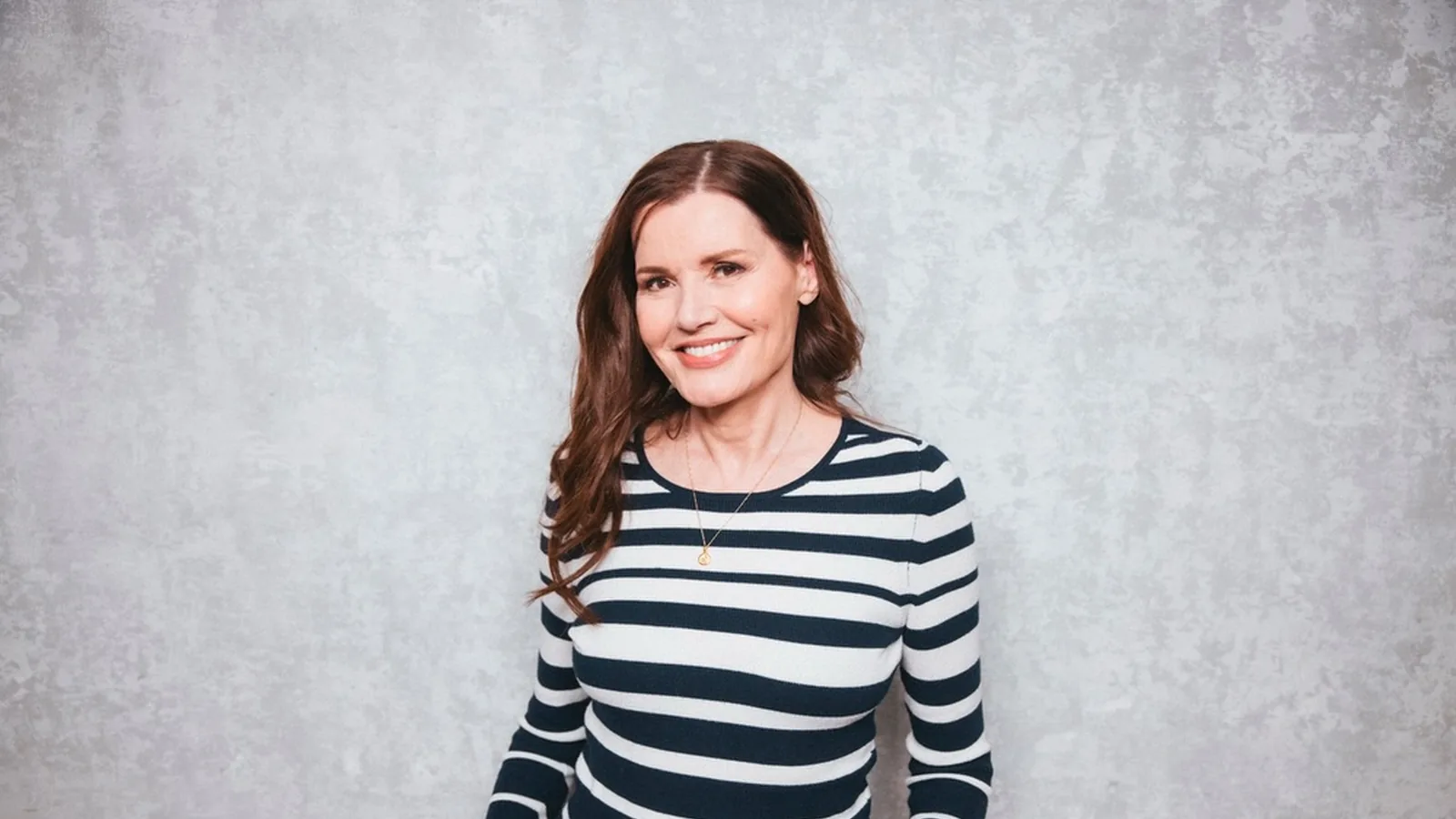 Geena Davis, de blusa listrada preta e branca, posa sorrindo diante de um fundo cinza
