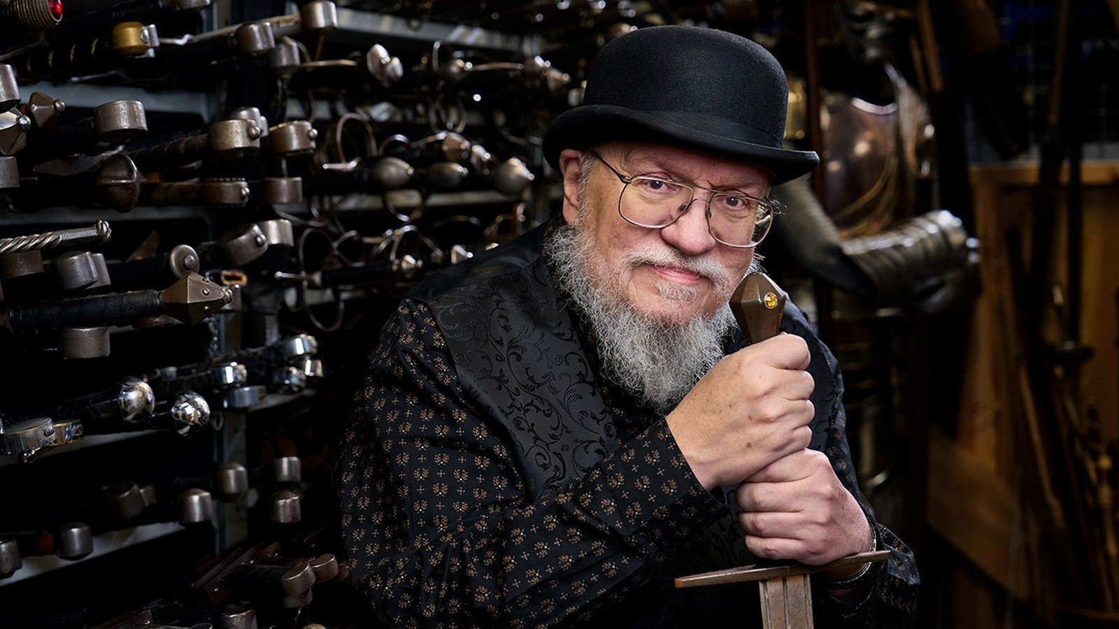 George R.R. Martin apoiado numa espada, cercado de armas de cenário