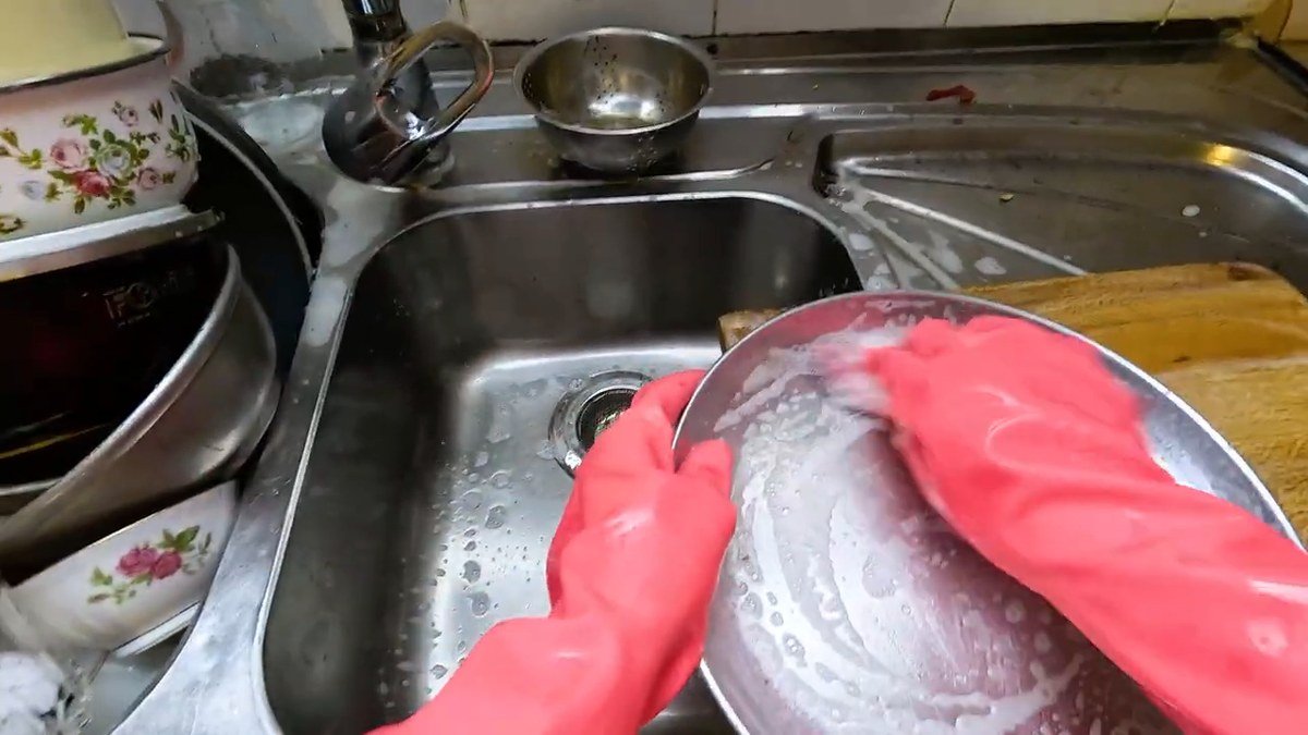 Mãos com luva rosa esfregando uma panela numa pia de aço cheia de louça, gravadas de cima em primeira pessoa