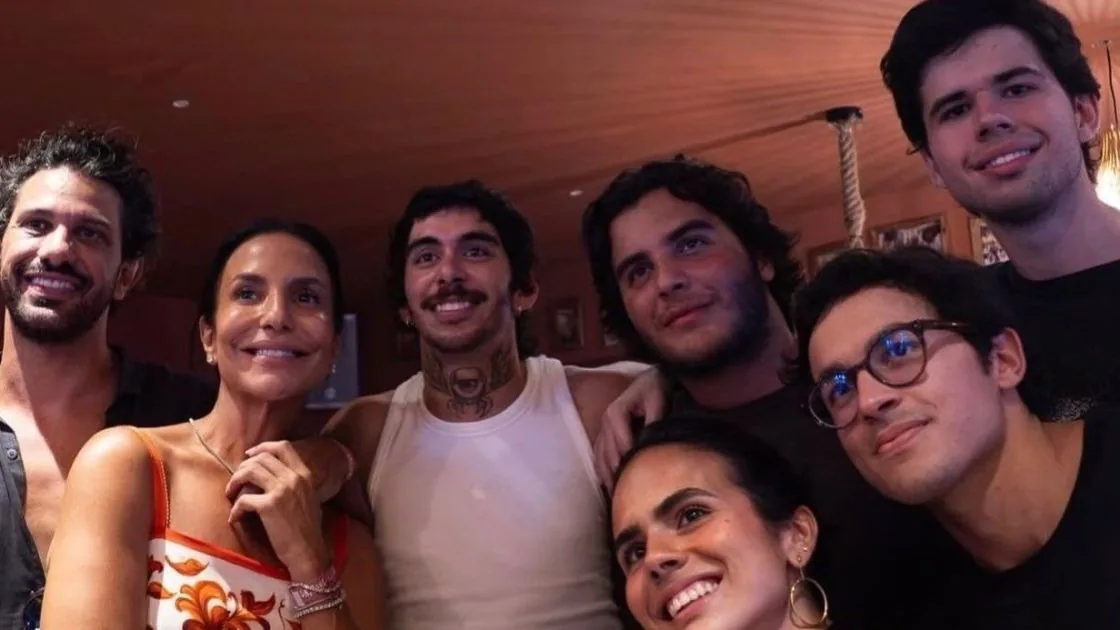 Ivete Sangalo sorrindo em meio a um grupo de sete pessoas numa festa, sob teto de madeira
