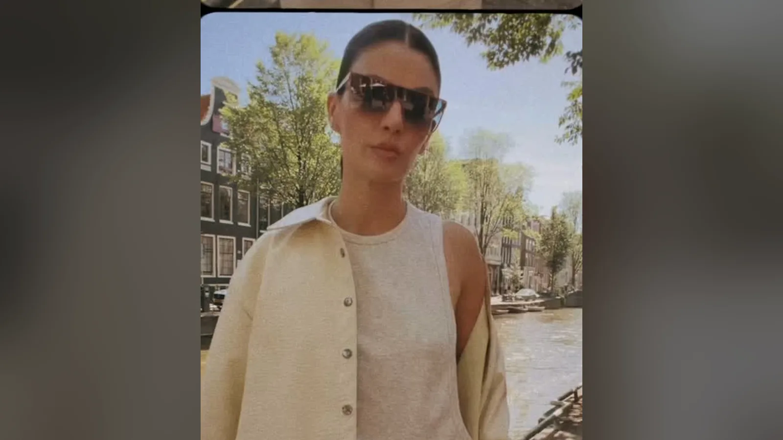 Monica Benini, de óculos escuros e cardigã claro, posa diante de um canal de Amsterdã
