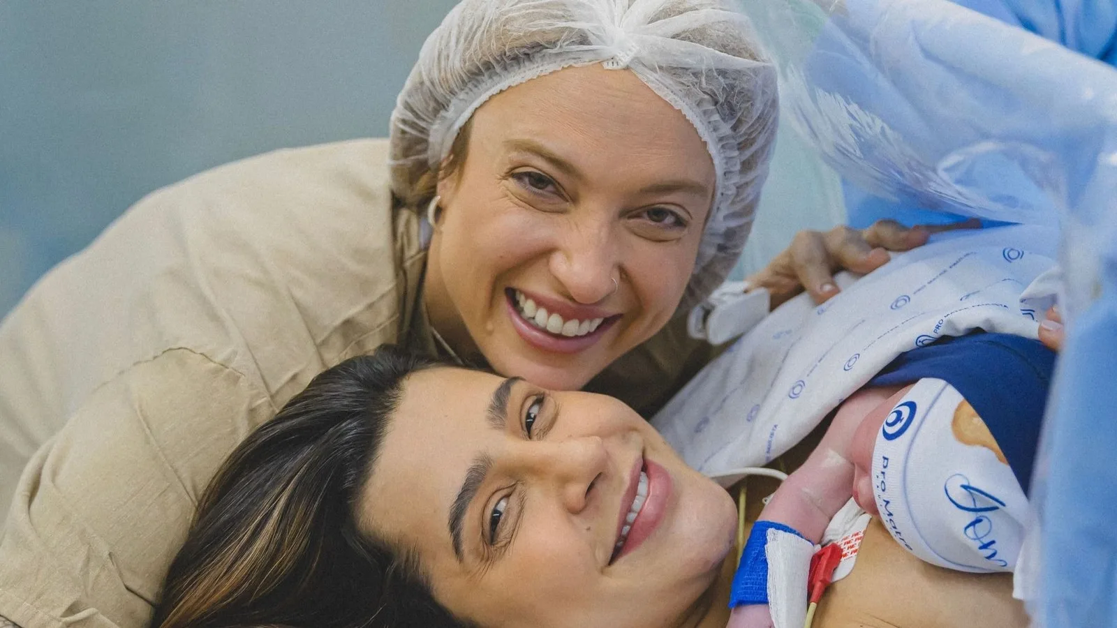 Lauana Prado deitada na sala de parto e Tati Dias de touca cirúrgica, as duas sorrindo, com o recém-nascido ao lado