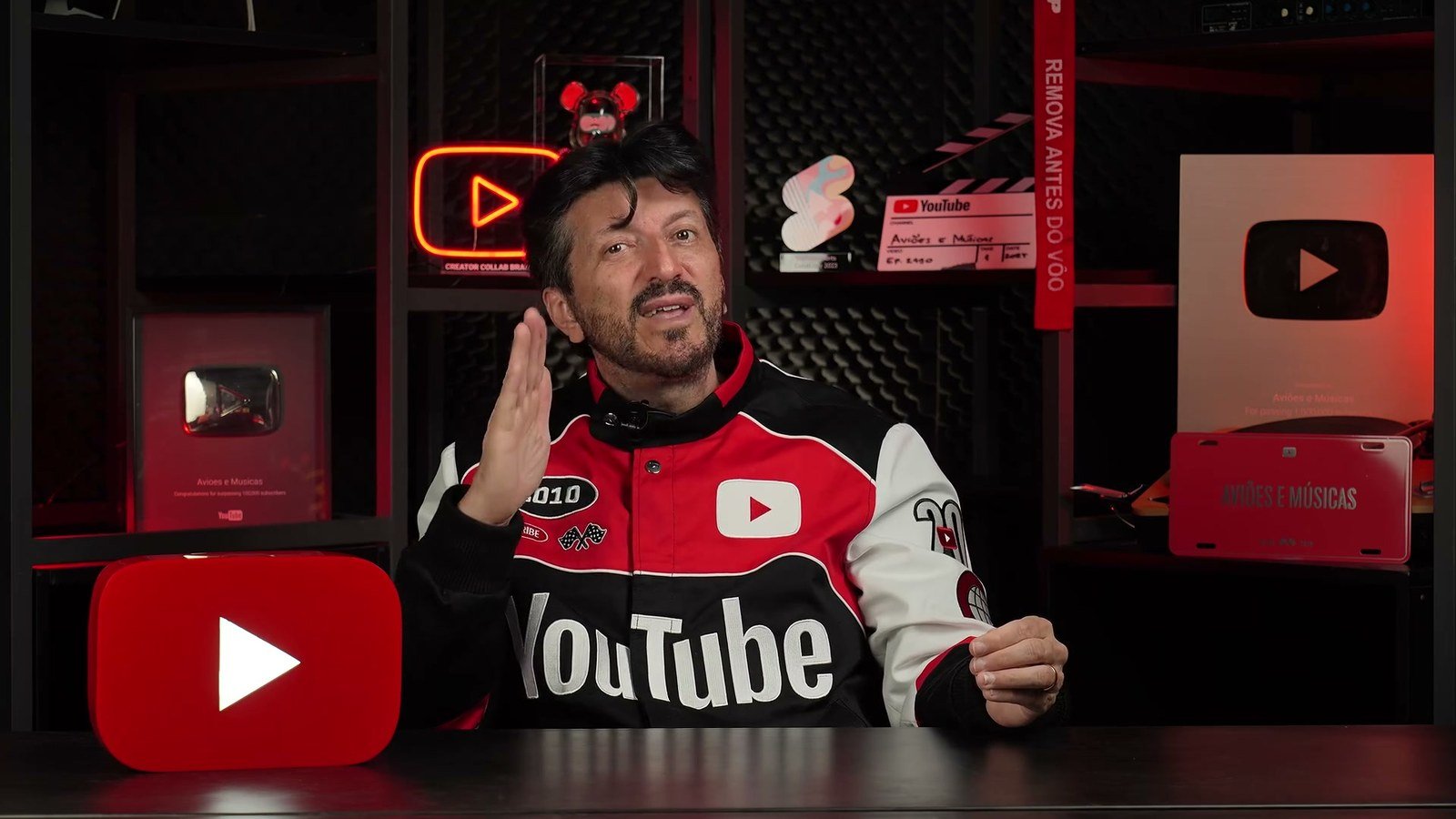 Lito Sousa no estudio do canal Avioes e Musicas, de jaqueta, cercado por placas do YouTube