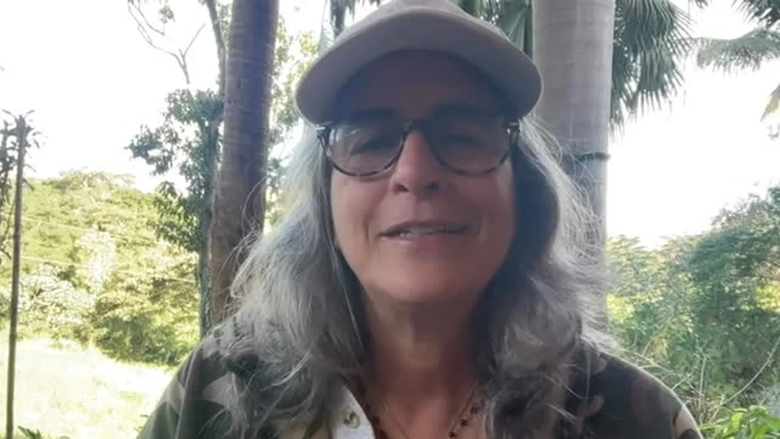 Lúcia Veríssimo, de boné e óculos, sorri ao ar livre com mata e palmeiras ao fundo