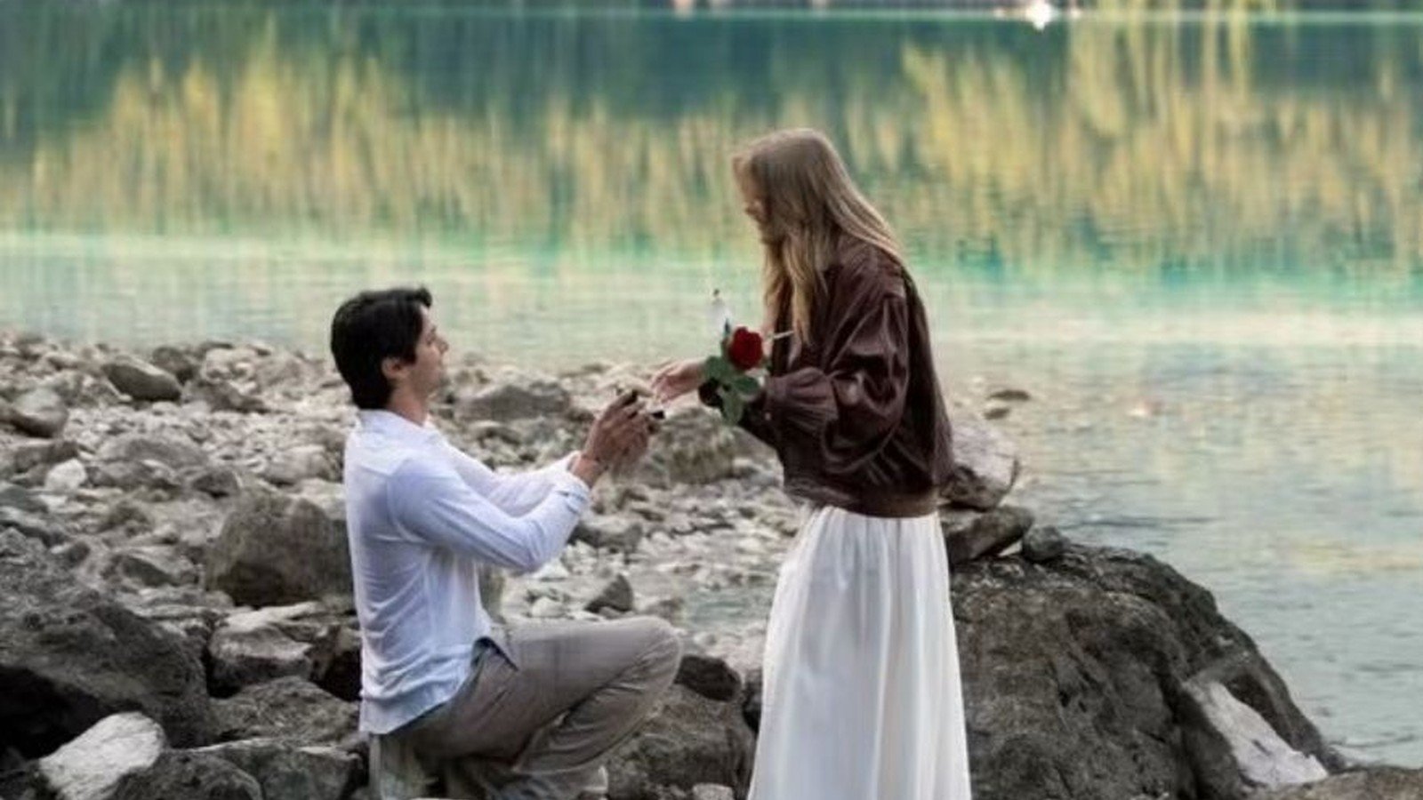 Luis Ribeirinho de joelho sobre as pedras do Eibsee, entregando uma rosa vermelha a Luisa Sonza