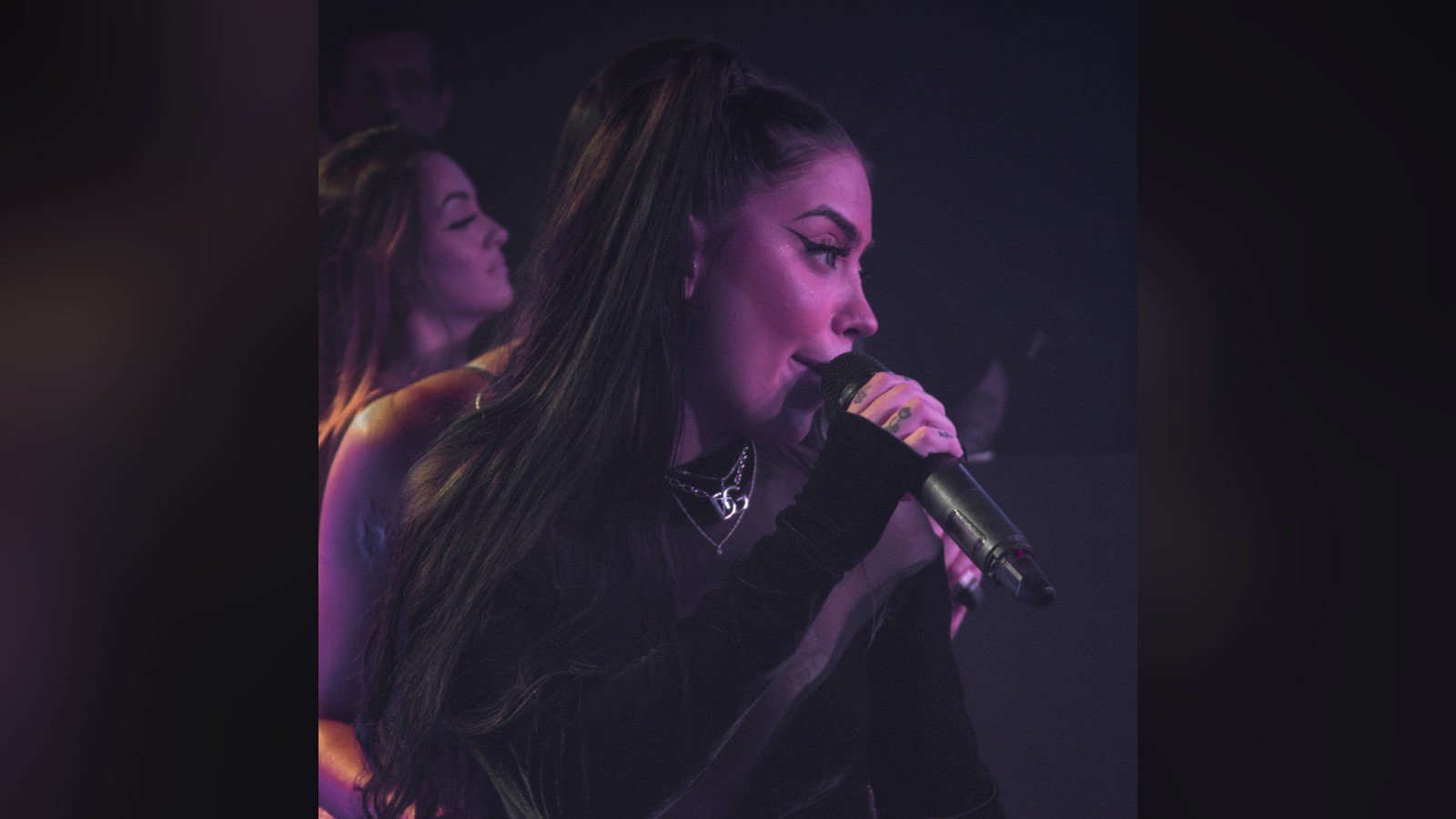MC Mirella cantando no palco, de microfone na mao, sob luz roxa