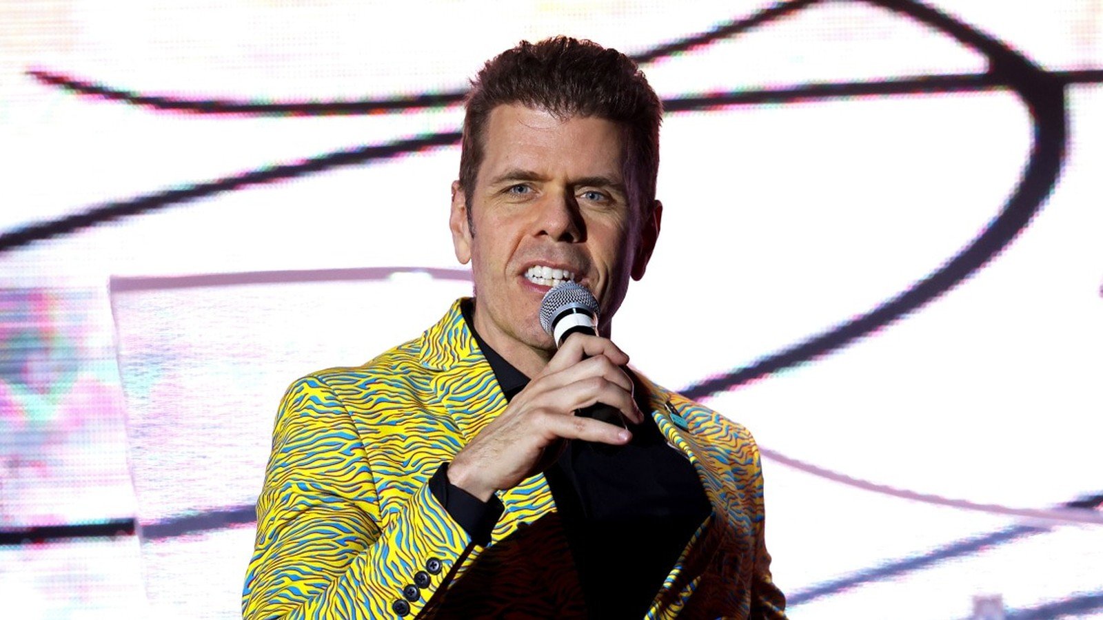 Perez Hilton fala ao microfone num palco iluminado