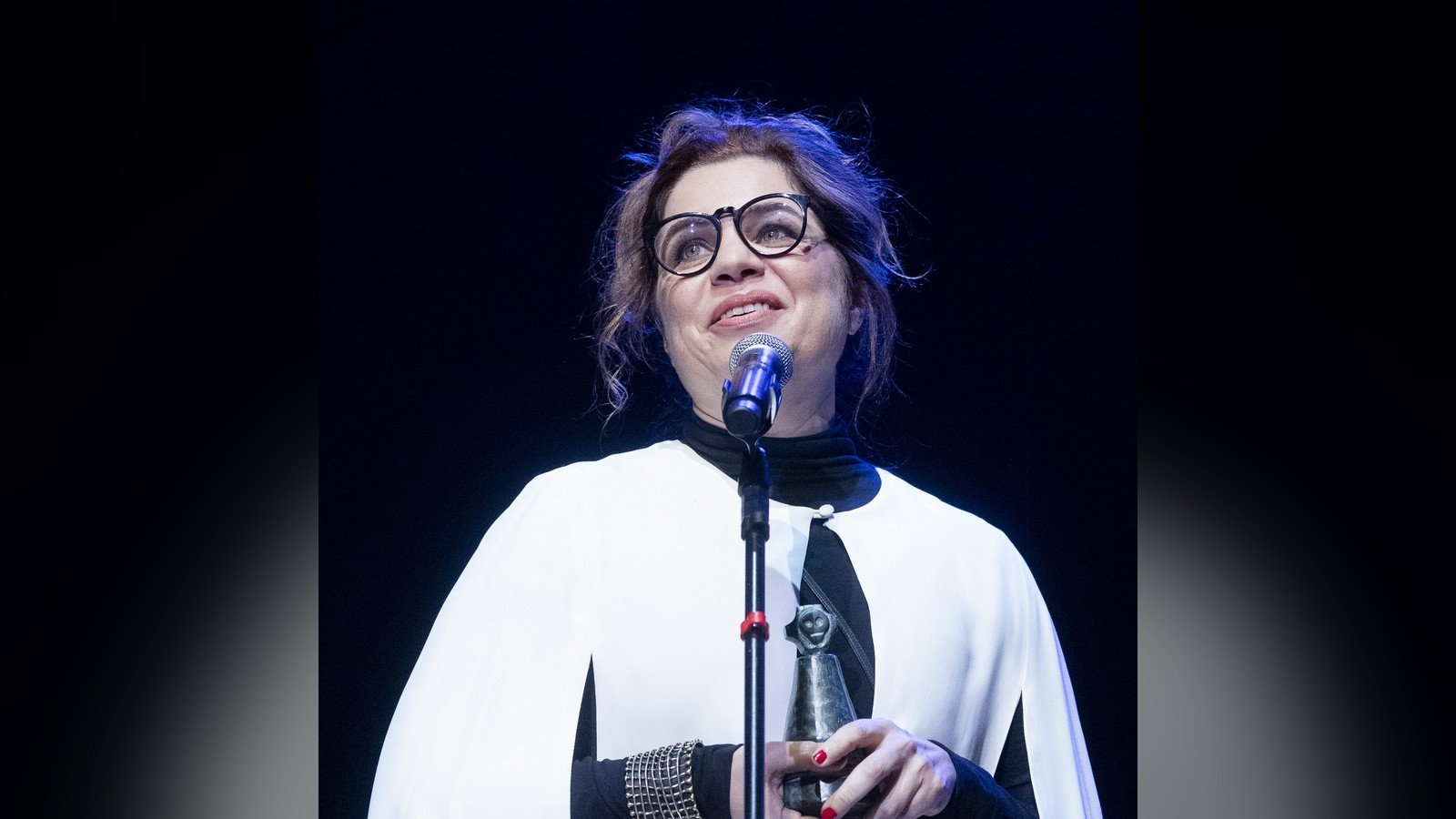 Isabel Teixeira de oculos e roupa branca, falando ao microfone em premiacao