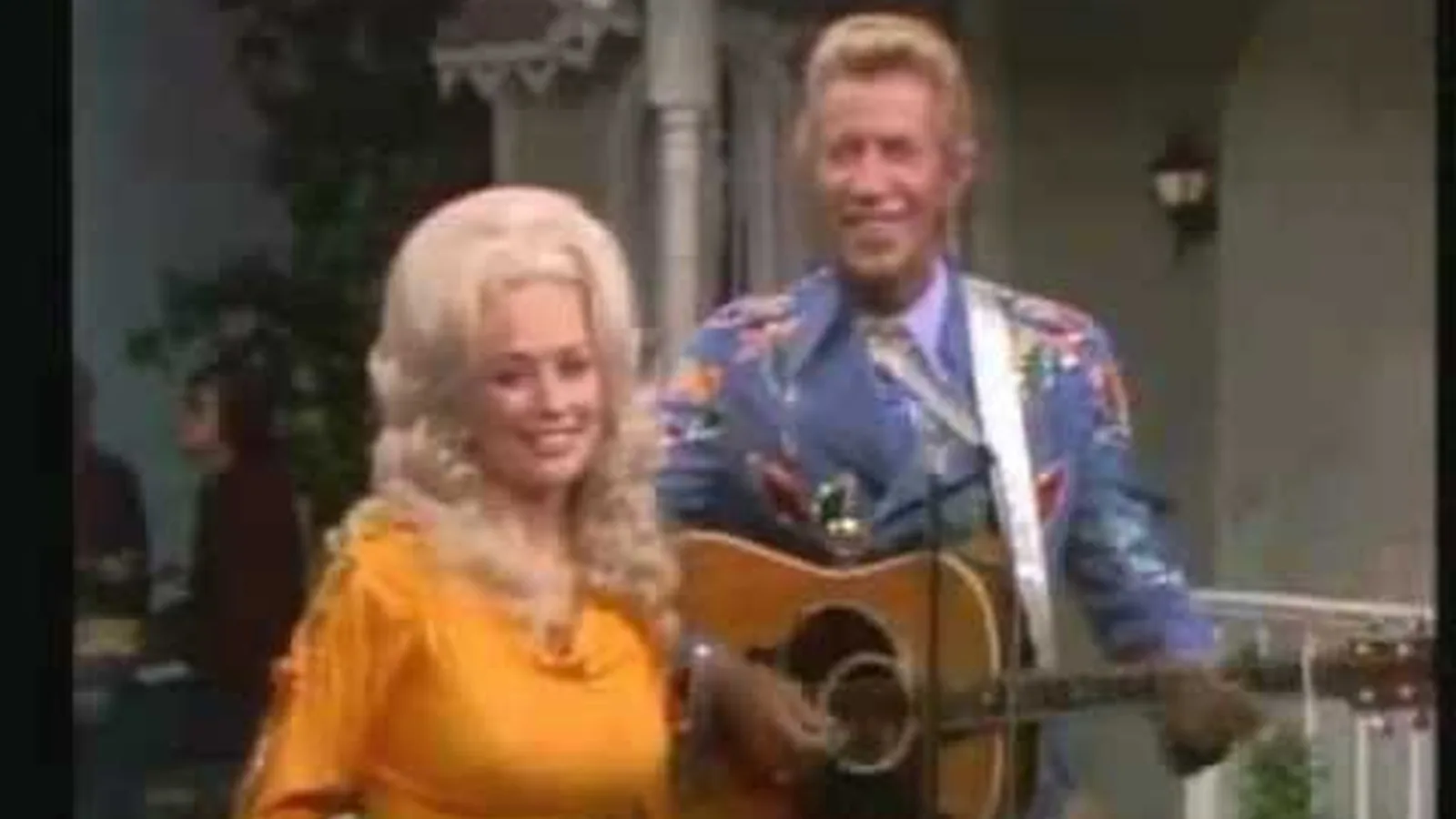 Dolly Parton, de vestido amarelo, ao lado de Porter Wagoner, que segura um violão, em cenário de programa de TV dos anos 1970