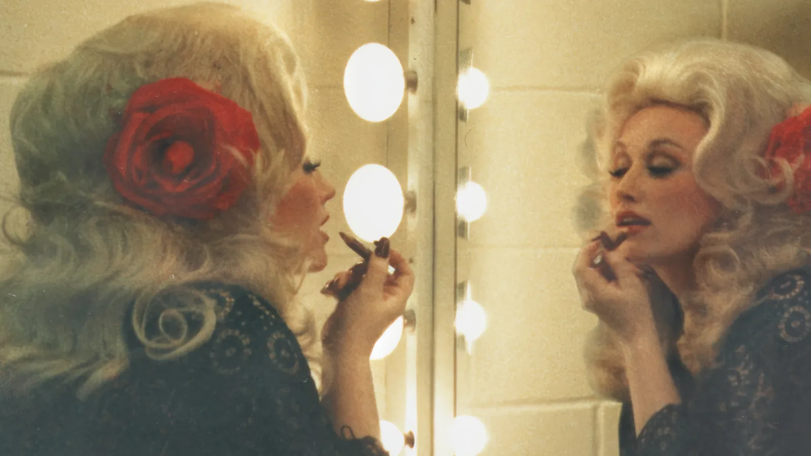 Dolly Parton se maquia diante do espelho iluminado de um camarim, com uma rosa vermelha no cabelo