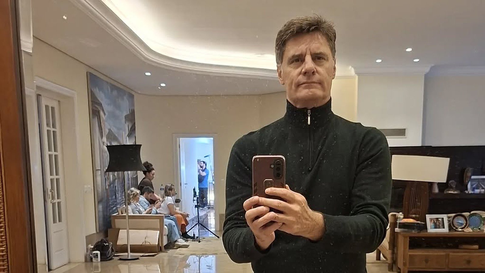 Renato Scarpin de preto, fotografando o próprio reflexo no espelho do saguão de um teatro