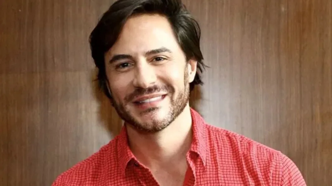 Ricardo Tozzi sorrindo, de camisa xadrez vermelha, com barba por fazer