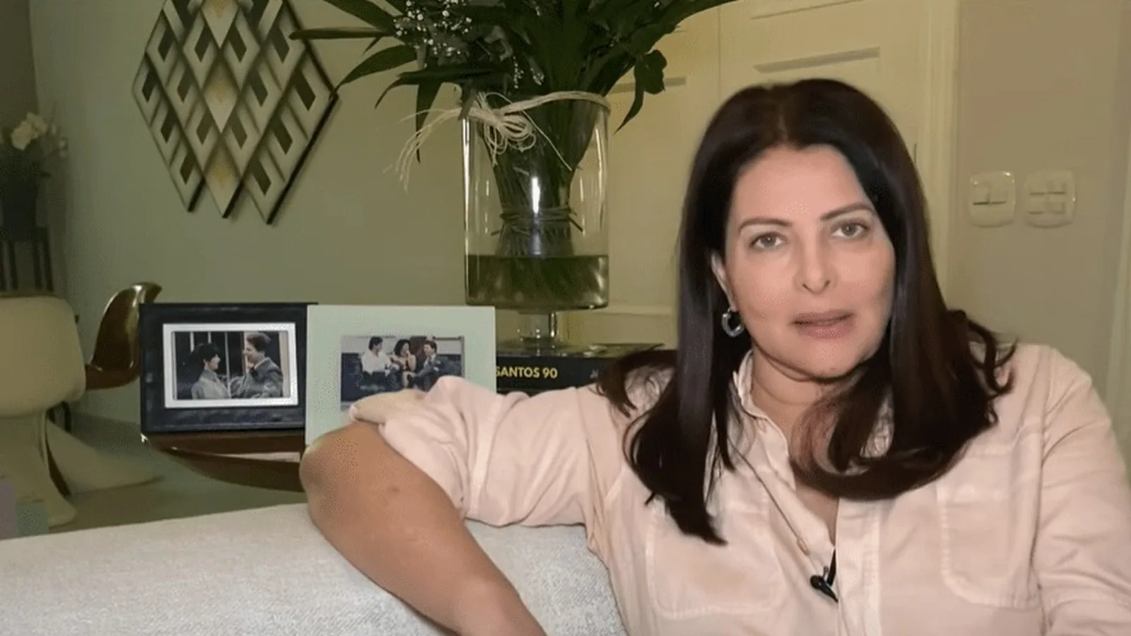 Sônia Lima sentada em casa durante entrevista à CNN Brasil, com porta-retratos ao fundo
