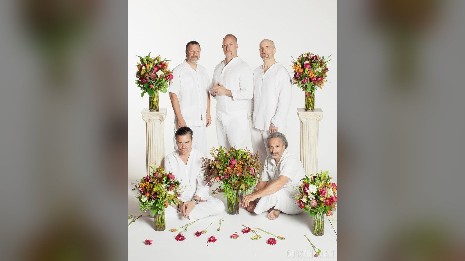 Os cinco integrantes do Faith No More posam vestidos de branco entre vasos de flores