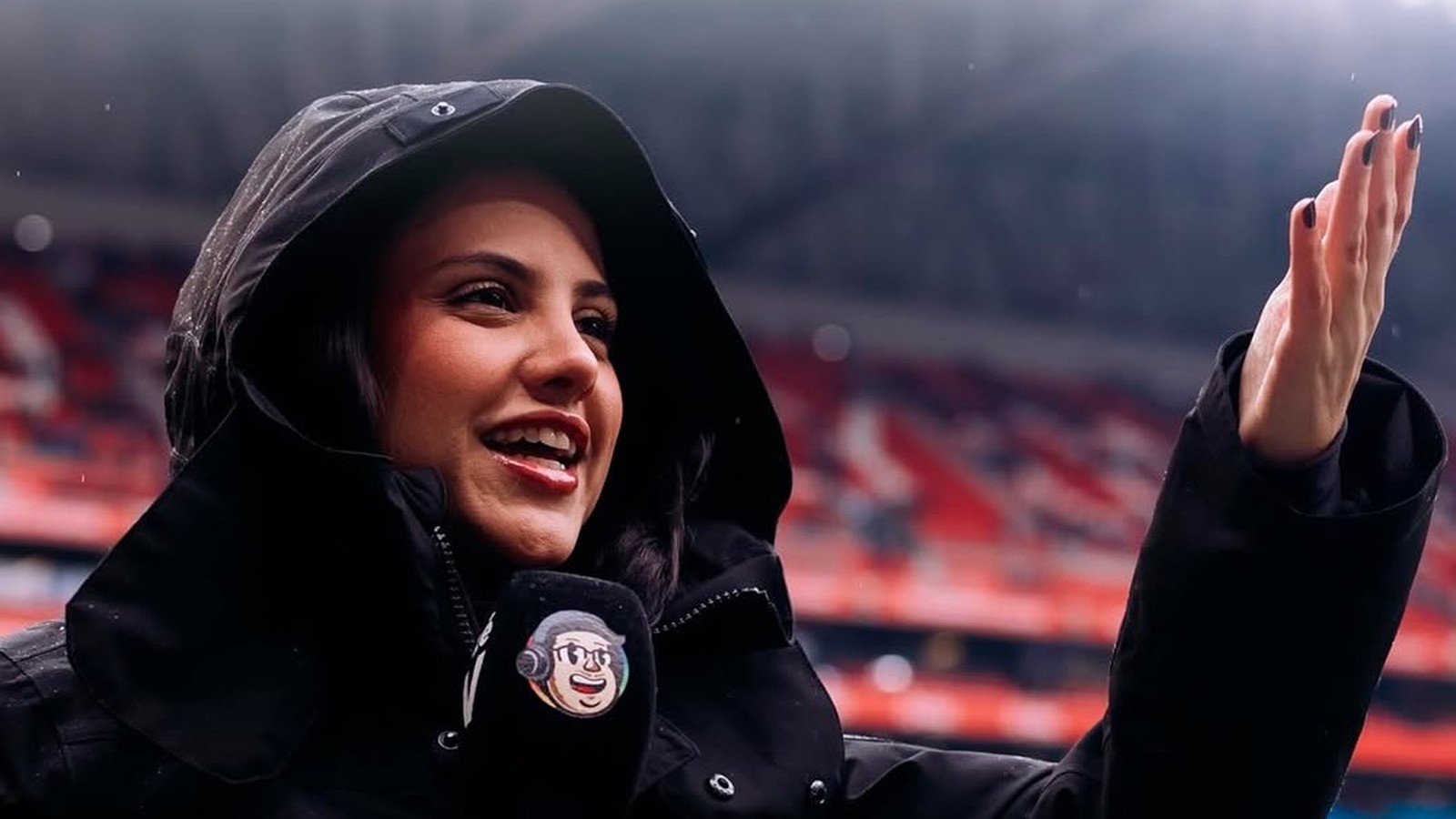 Victoria Leite de capuz e casaco preto, sorrindo com o microfone na mao dentro de um estadio