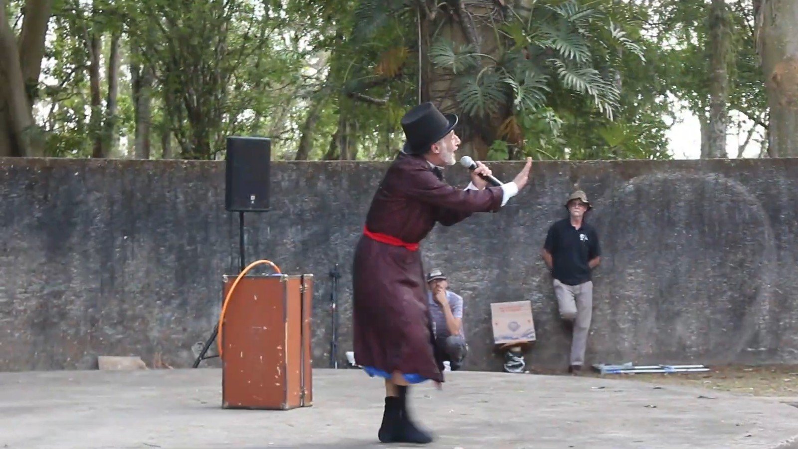 Carlos Cesare de roupao vinho, chapeu coco e gravata borboleta durante numero de palhaco, com mala de madeira ao lado