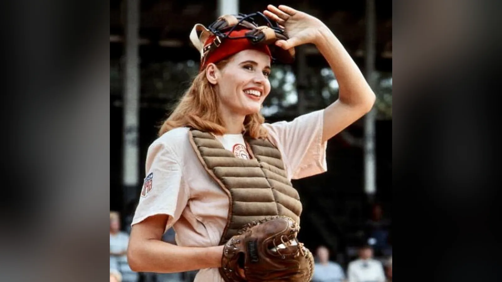 Geena Davis vestida de jogadora de beisebol, com máscara de receptora na cabeça e luva na mão
