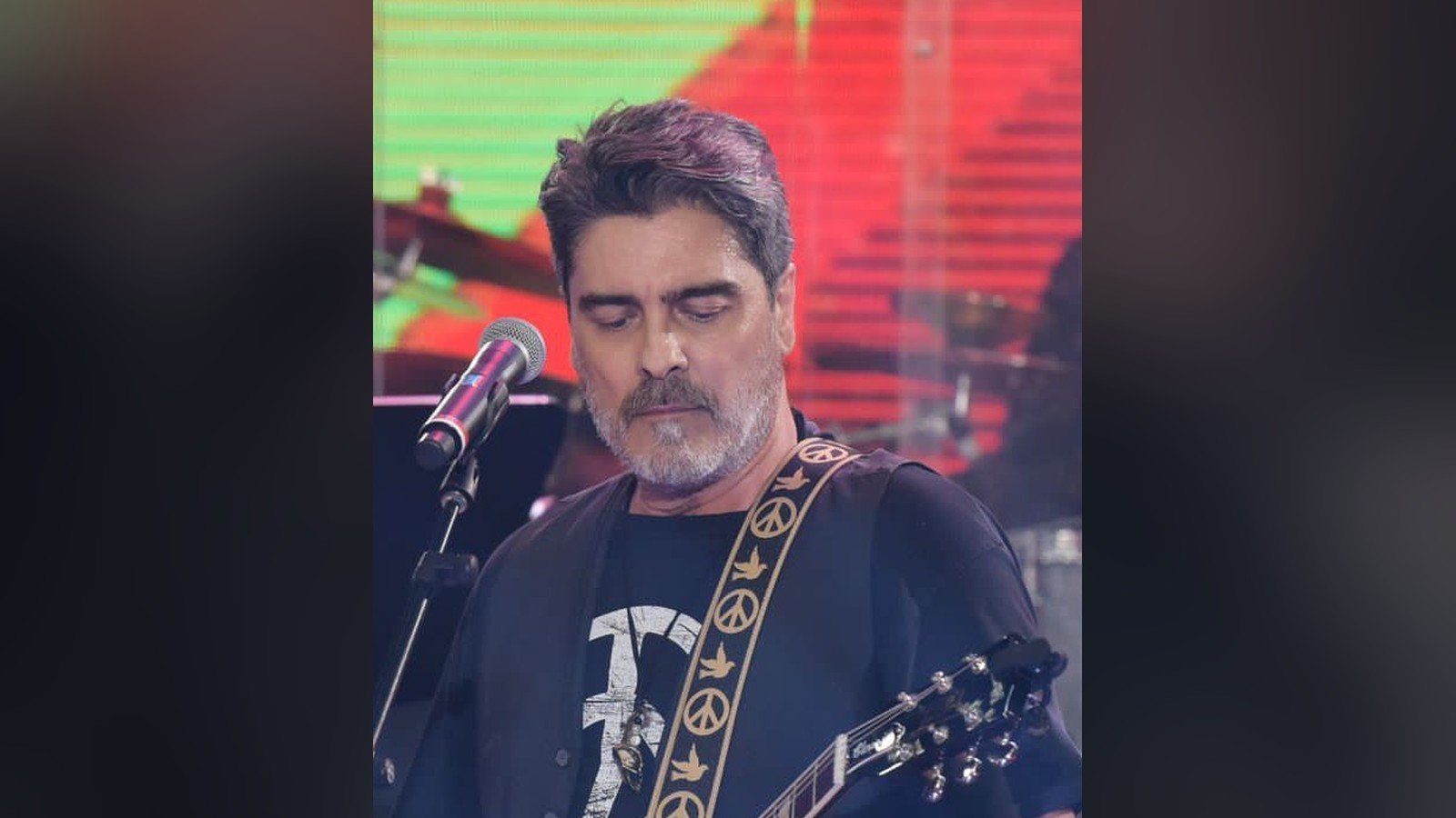 Junno Andrade toca guitarra diante do microfone num palco iluminado de verde e vermelho