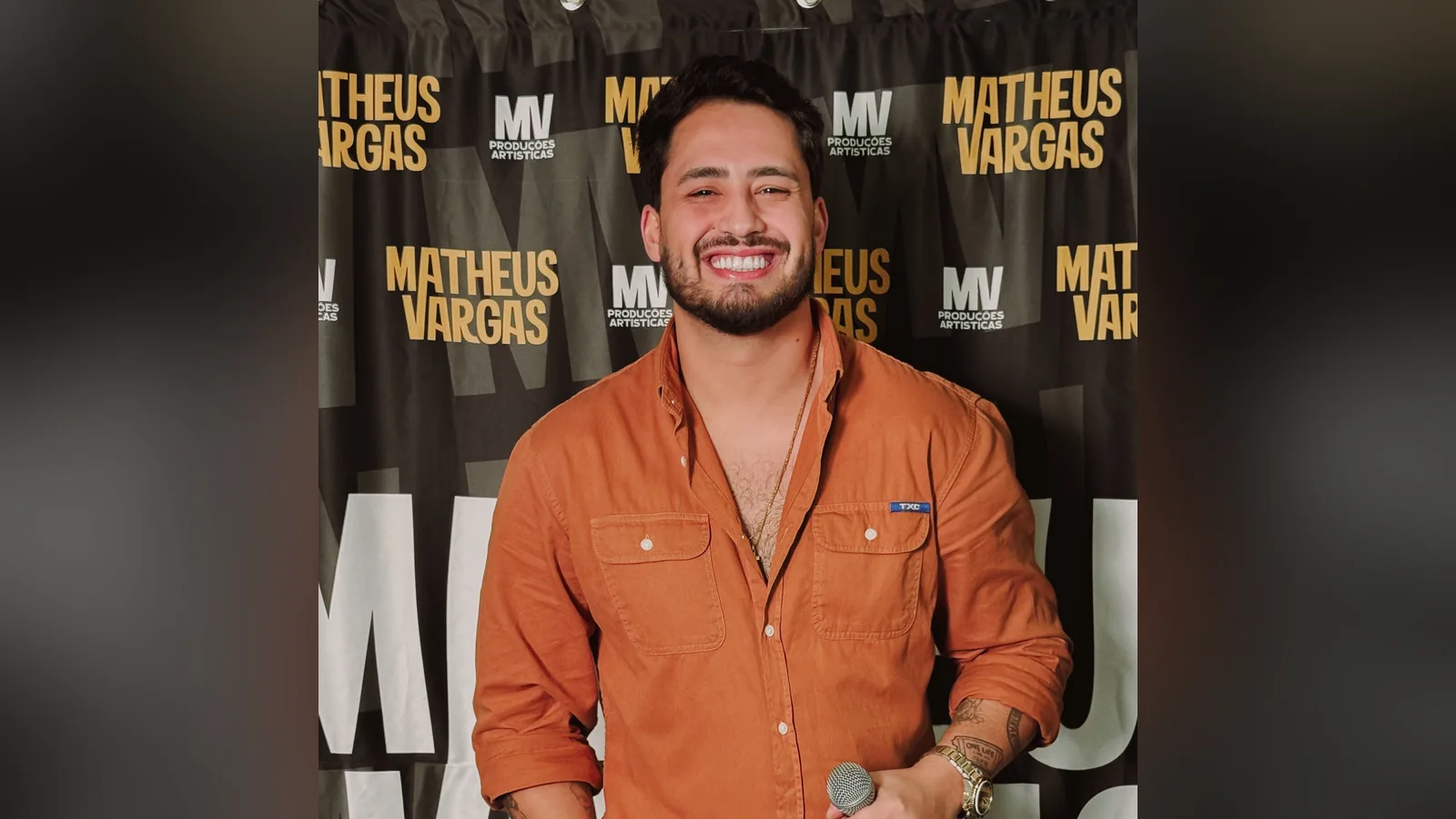 Matheus Vargas de camisa laranja e jeans, sorrindo com o microfone na mão, diante de painel com o nome dele repetido