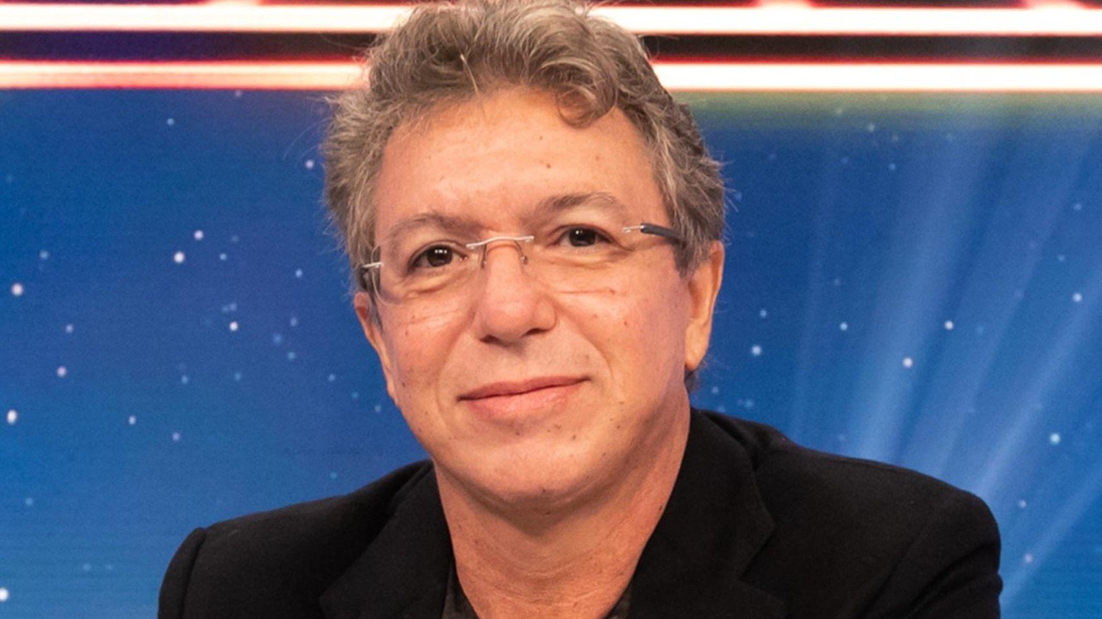Boninho de terno escuro, sorrindo, em cenario azul de estudio