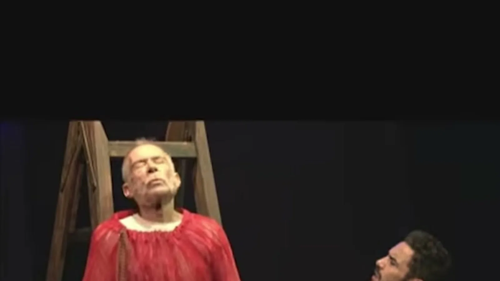 Ricardo Blat, de figurino vermelho, em cima de uma escada no palco, com Caio Blat de costas em cena da peça Subversão Kafka