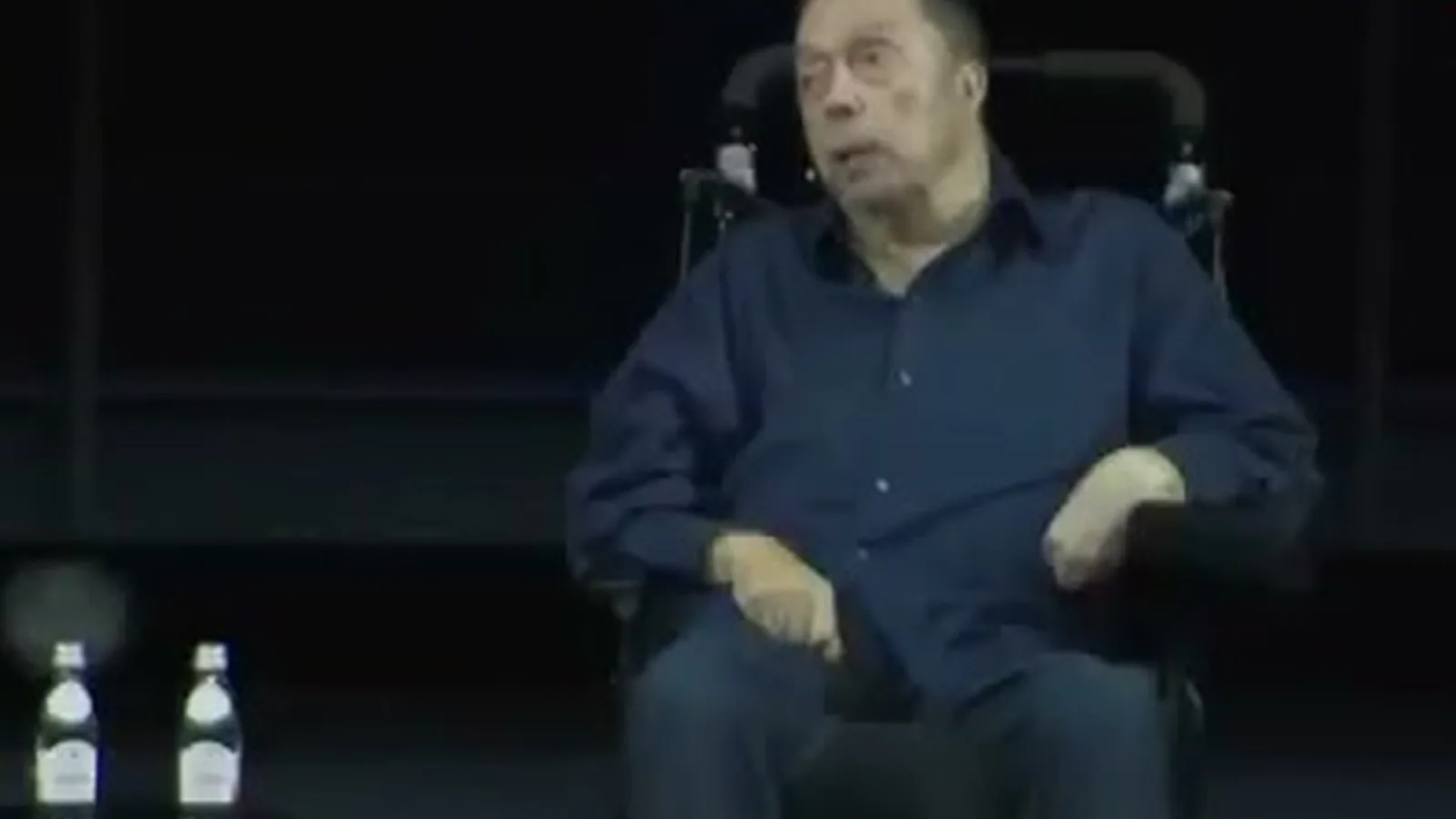 Tim Curry, de camisa azul e cabelo grisalho, sentado em cadeira de rodas motorizada no palco escuro do Academy Museum