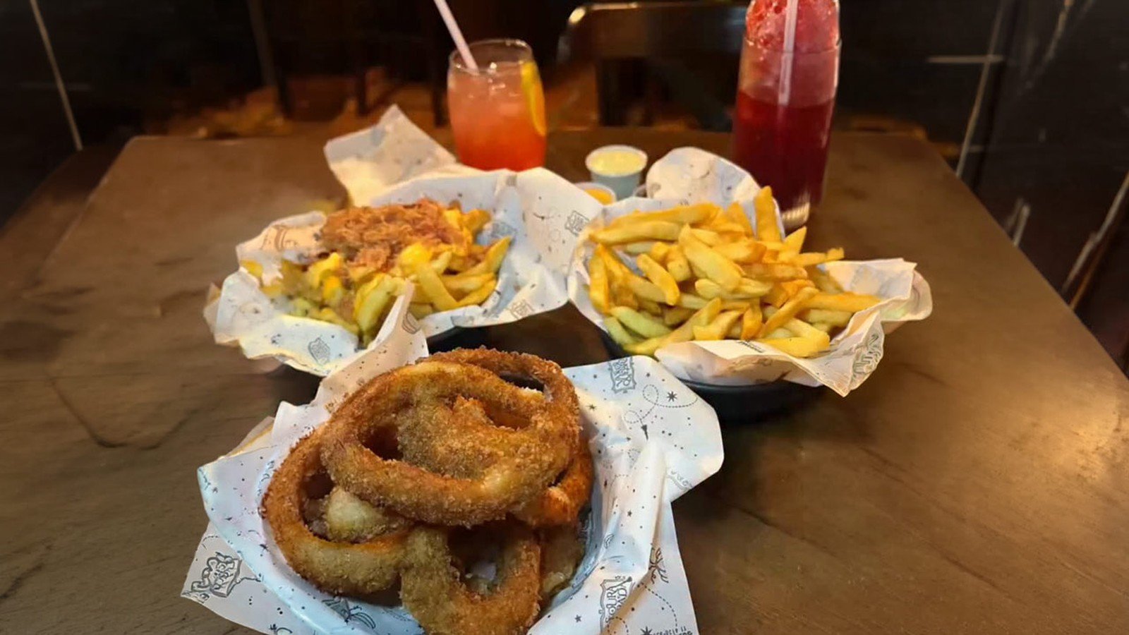 Porções de batata frita, anéis de cebola empanados e duas bebidas coloridas sobre mesa de madeira