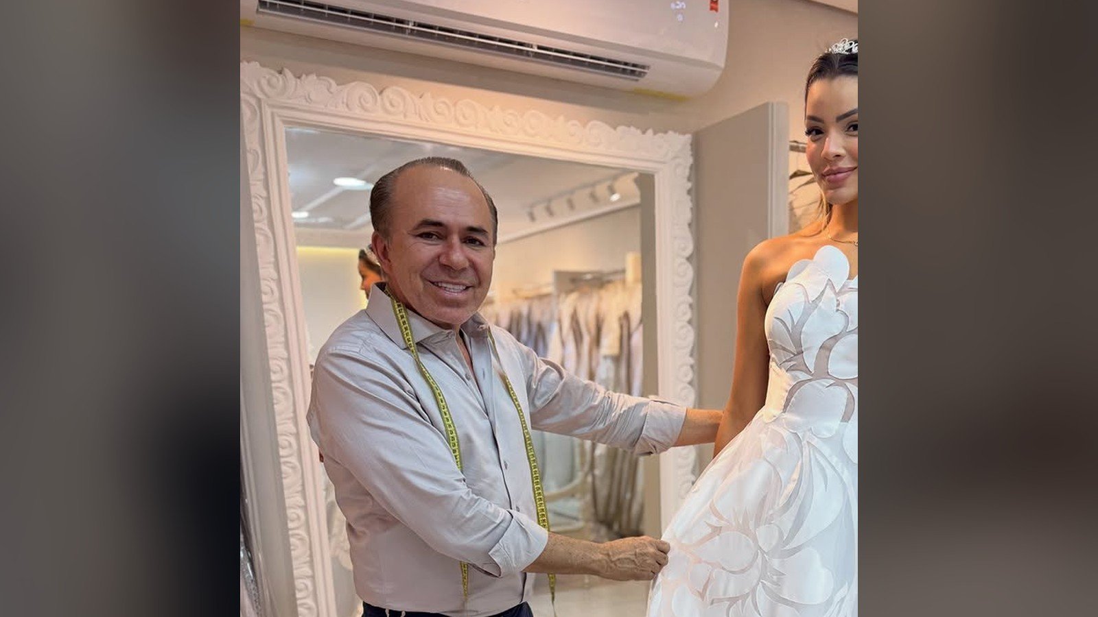 Emilly Araujo prova um vestido de noiva branco em um atelie, ao lado de um homem