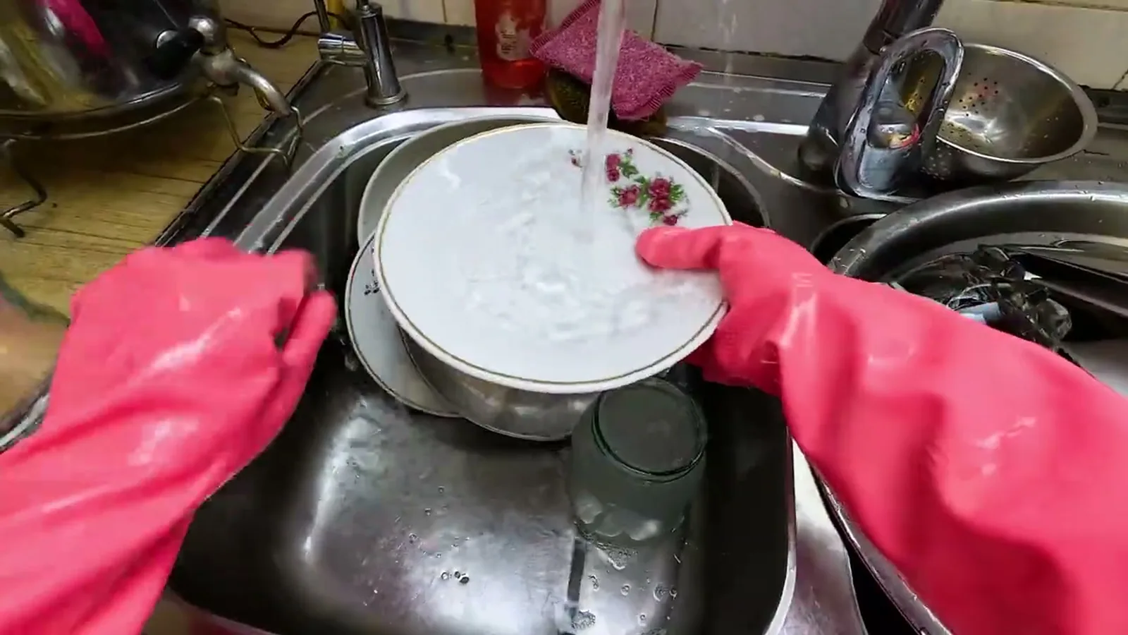 Mãos com luva rosa lavando um prato sob a torneira, gravadas de cima pelo celular preso à cabeça