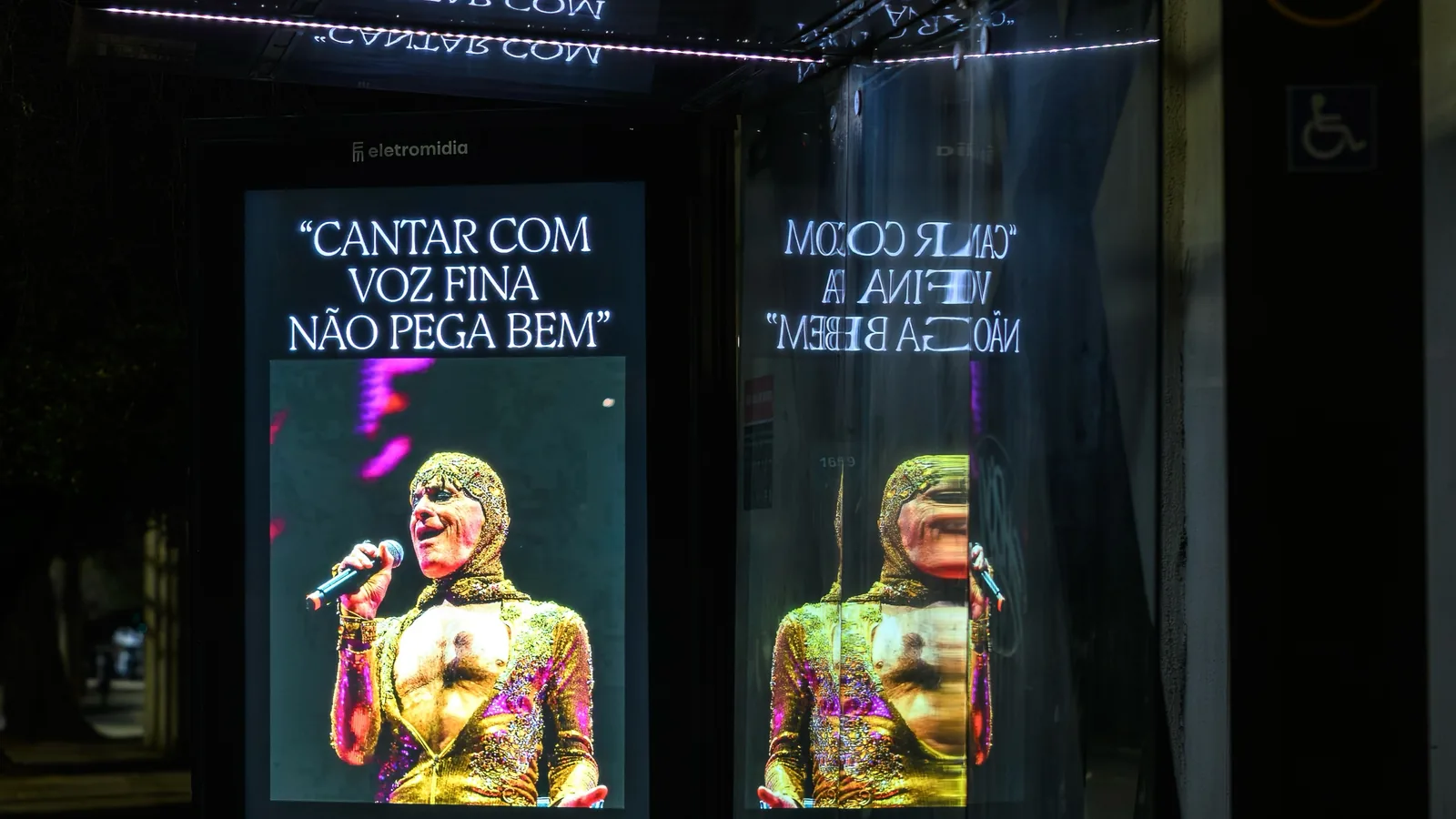 Painel iluminado em ponto de ônibus com a frase Cantar com voz fina não pega bem e foto de Ney Matogrosso cantando