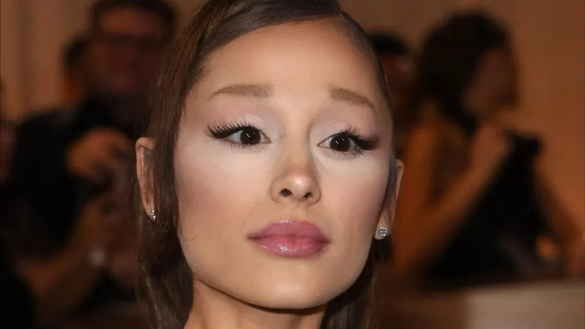Ariana Grande em close, cabelo preso, cercada por fotógrafos desfocados ao fundo