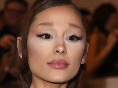 Ariana Grande em close, cabelo preso, cercada por fotógrafos desfocados ao fundo
