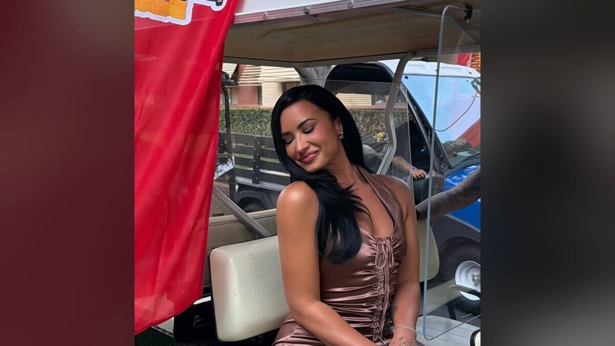Demi Lovato sentada em um carrinho de golfe, sorrindo, diante de um banner do Camp Rock