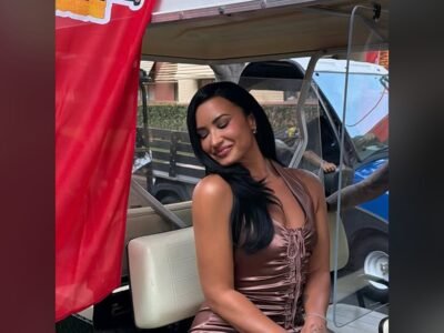 Demi Lovato sentada em um carrinho de golfe, sorrindo, diante de um banner do Camp Rock