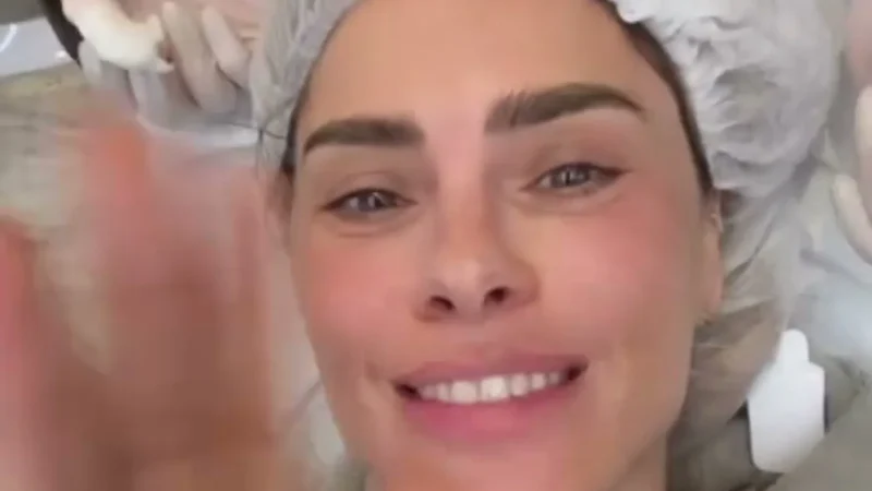 Carolina Dieckmann sorrindo, de touca cirúrgica, deitada na maca da clínica