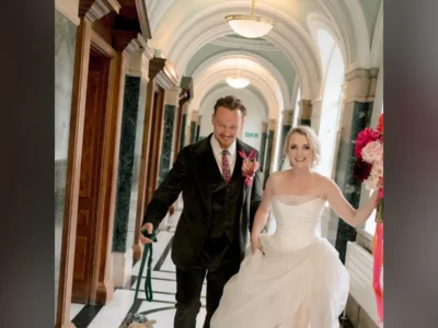 Evanna Lynch de vestido de noiva ao lado de Denis Skinner, de terno verde, num corredor com arcos