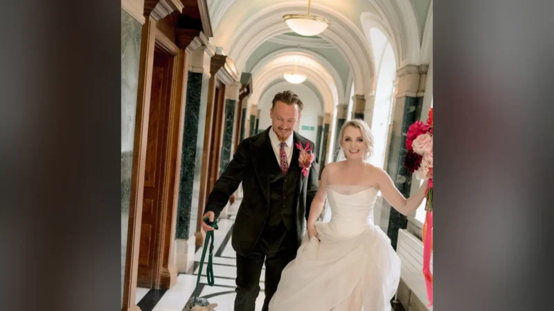 Evanna Lynch de vestido de noiva ao lado de Denis Skinner, de terno verde, num corredor com arcos