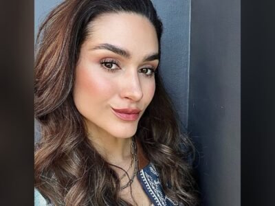 Fernanda Machado em close, cabelo castanho ondulado solto, encostada em uma parede cinza