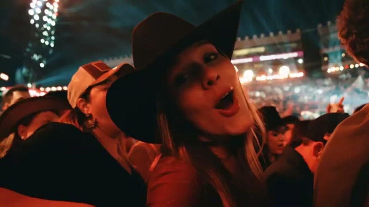Mulher de chapeu de peao canta no meio do publico da Festa do Peao de Barretos, com a arena iluminada ao fundo